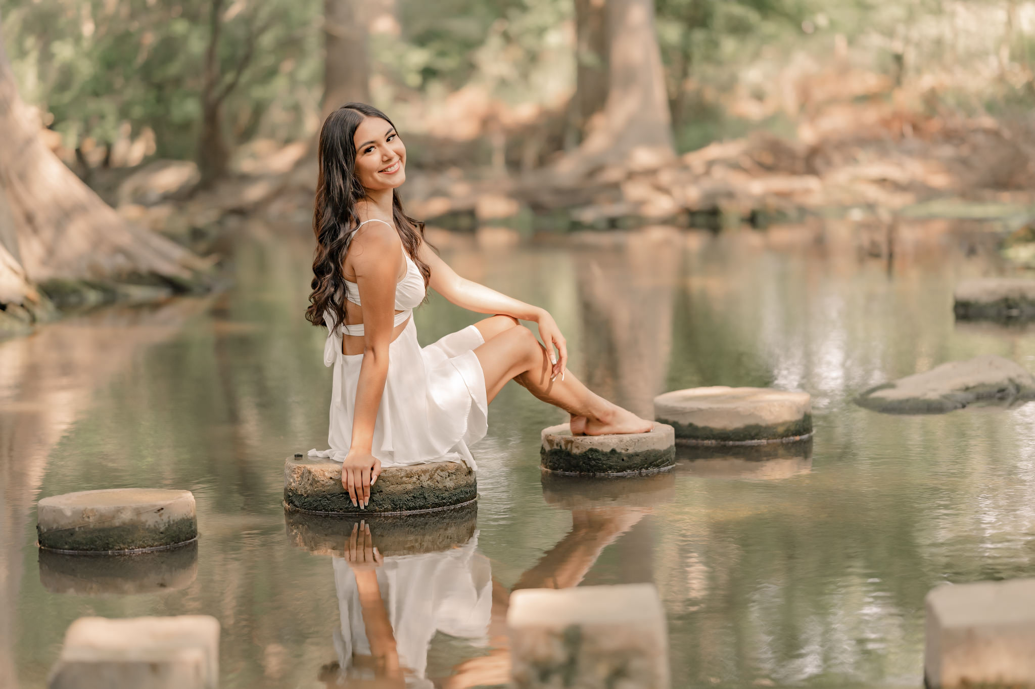 A young woman in a white dress sits on the stepping stones in the middle of the river at Cibolo Nature Center in Boerne, TX. 