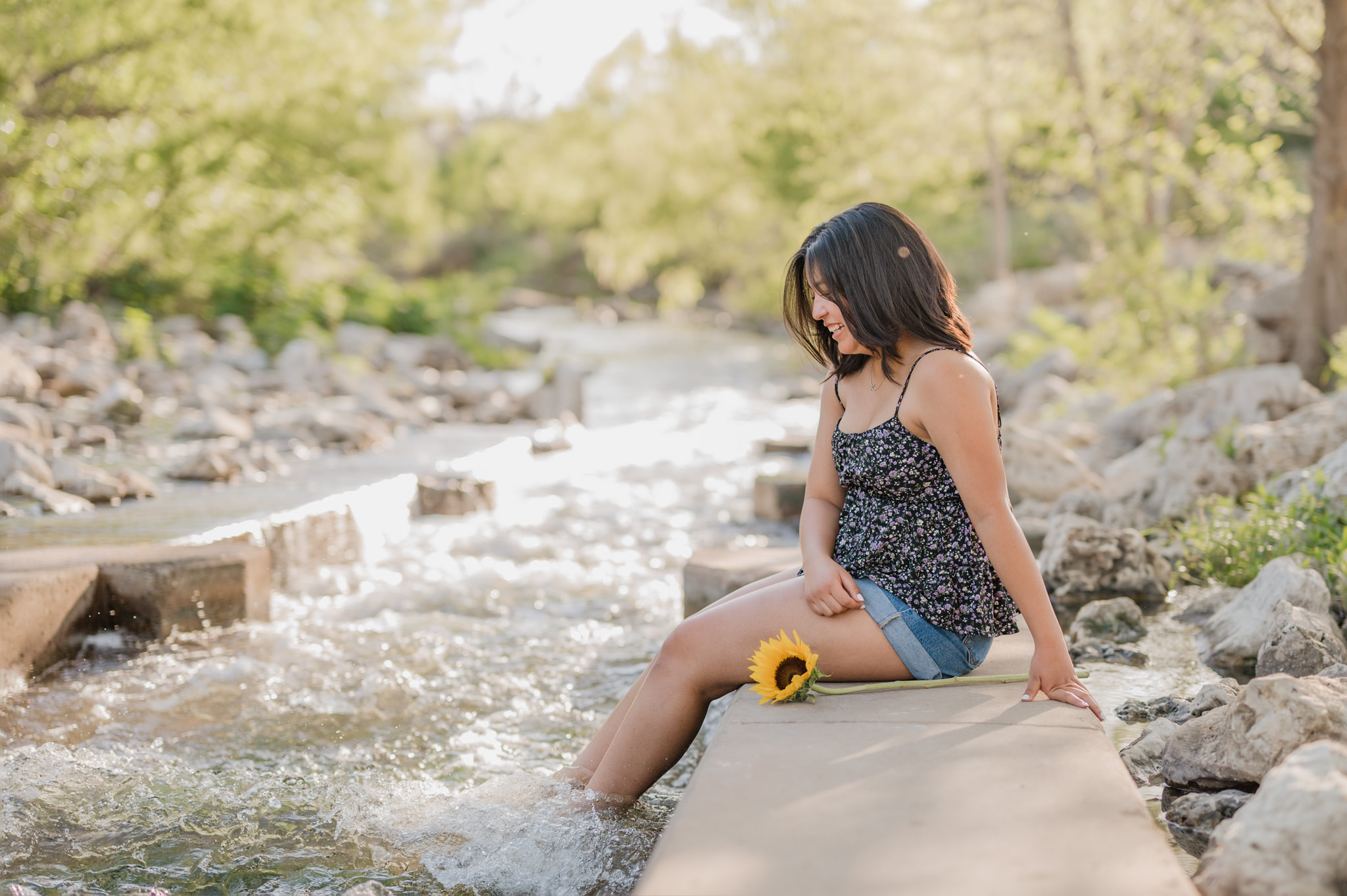A teenage girl sits on the concrete canoe chute at the river's edge at Confluence Park in San Antonio. Her feet are in the rushing water and a sunflower sits by her. 
