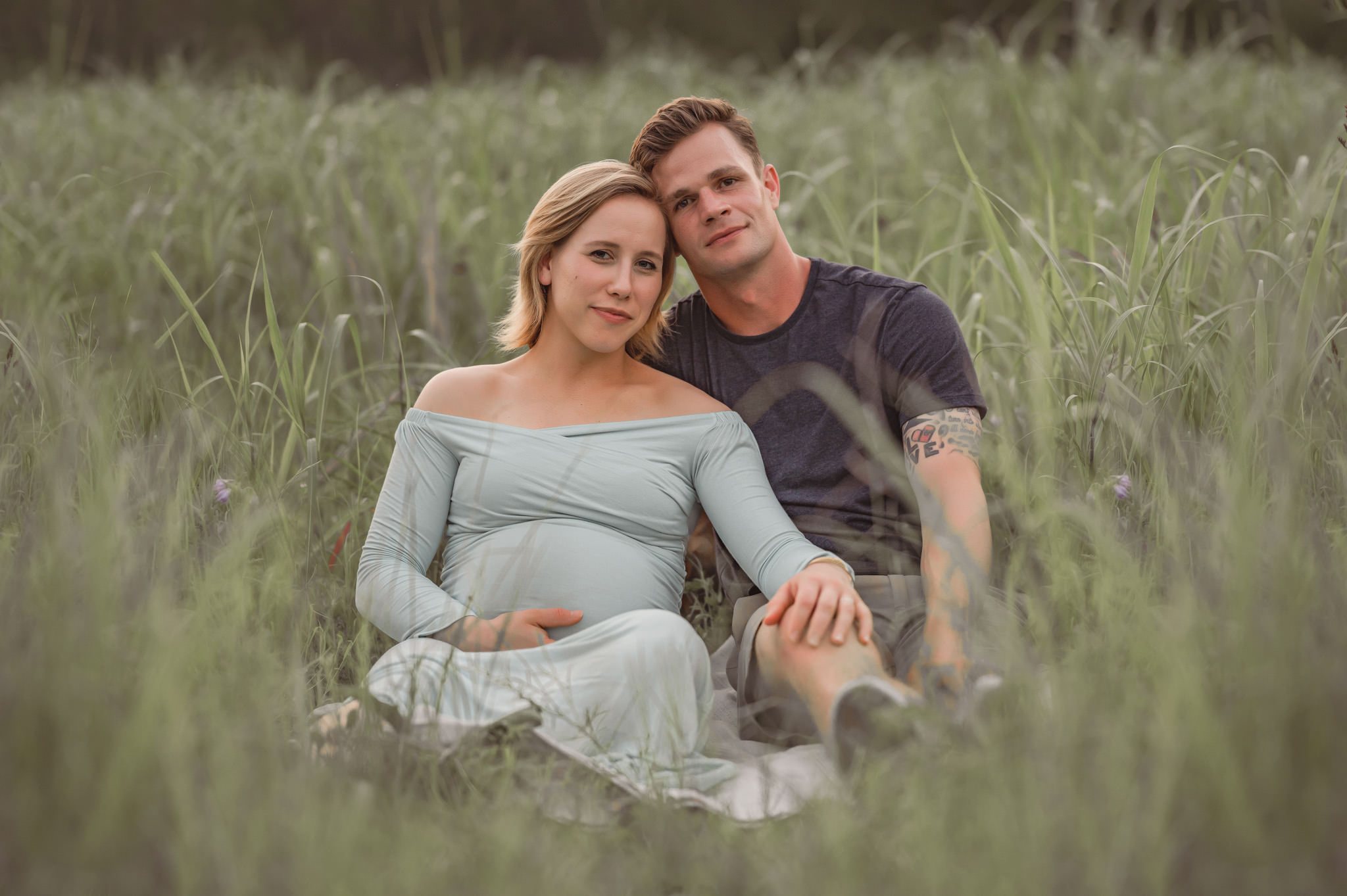 A young expecting couple sits and leans their heads together in a grass field at Crescent Bend Nature Park. 