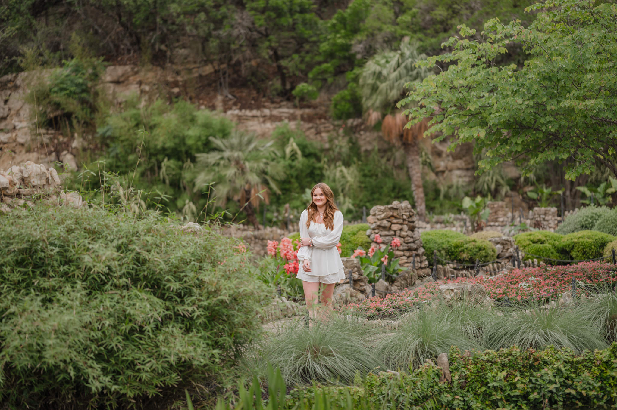 Pull-back landscape image of a senior girl in a white dress at the Japanese Tea Garden in San Antonio. She is surrounded by lush greenery.