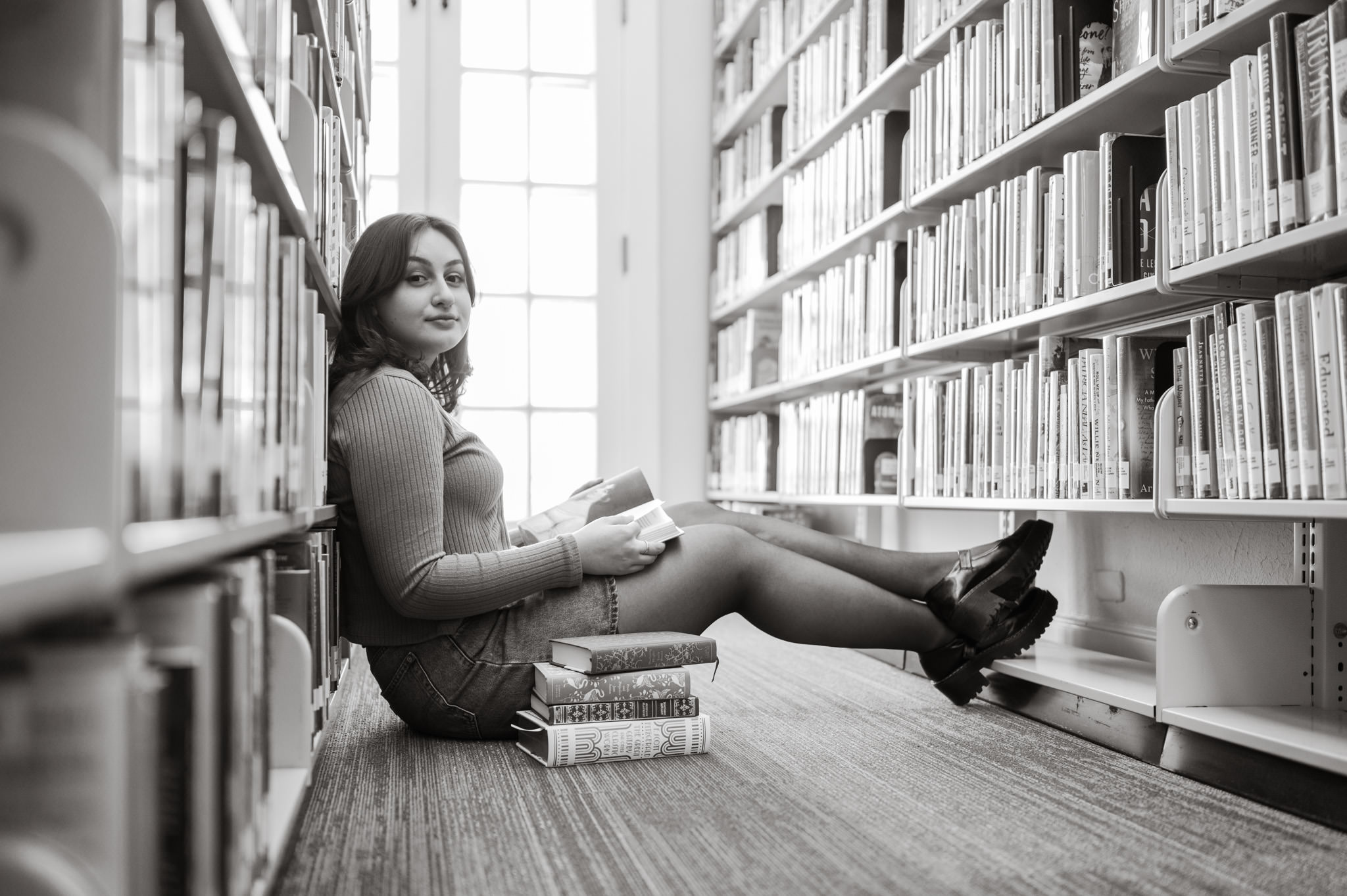 Black and white portrait of a girl sitting against a wall of books inside Landa Library. She has a stack of classic novels next to her and has one open on her lap. 