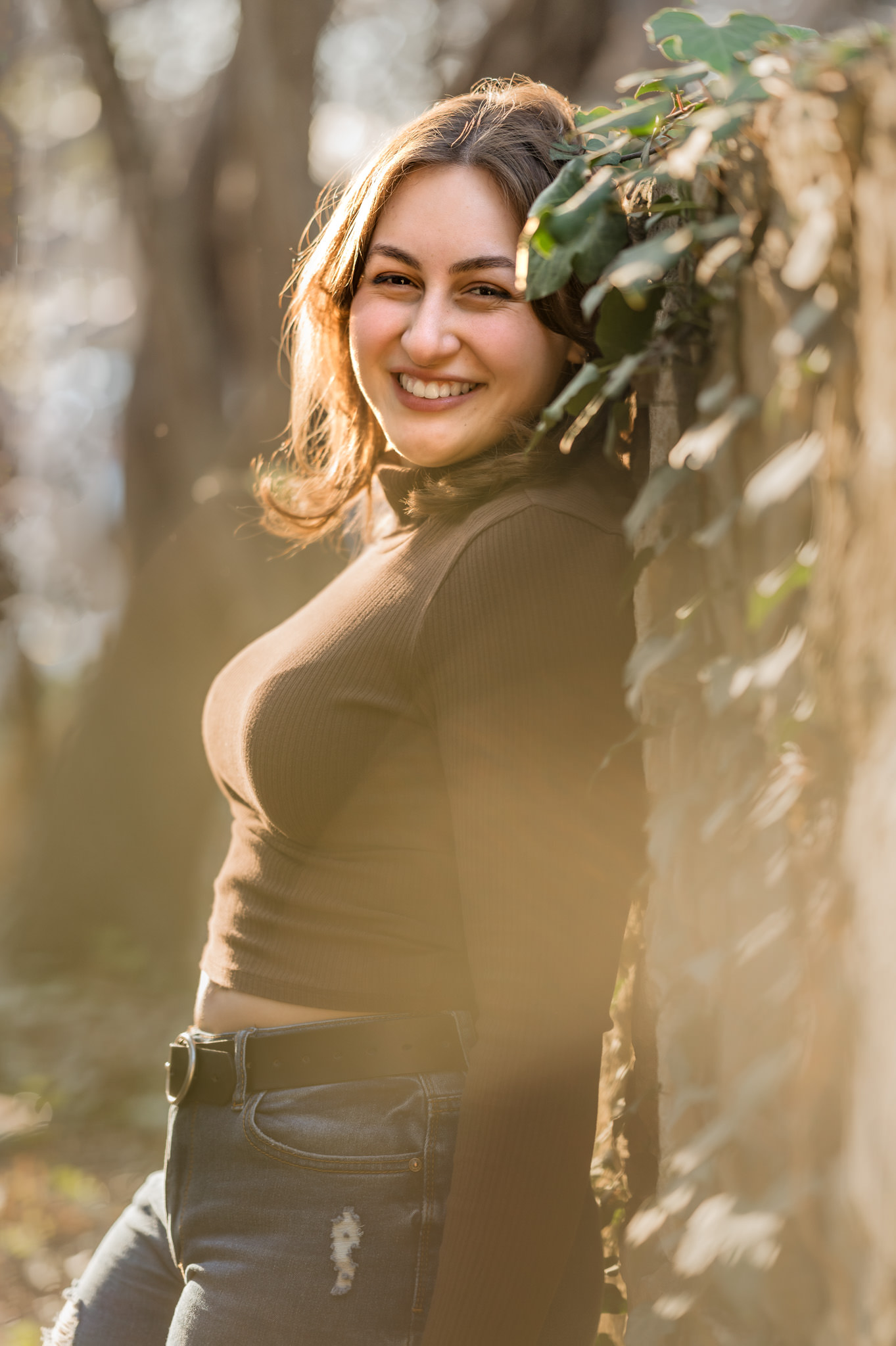 Senior portrait of a girl in jeans and a brown sweater leaning up against an ivy-covered wall on the grounds of the Landa Gardens Conservancy at Landa Library. 