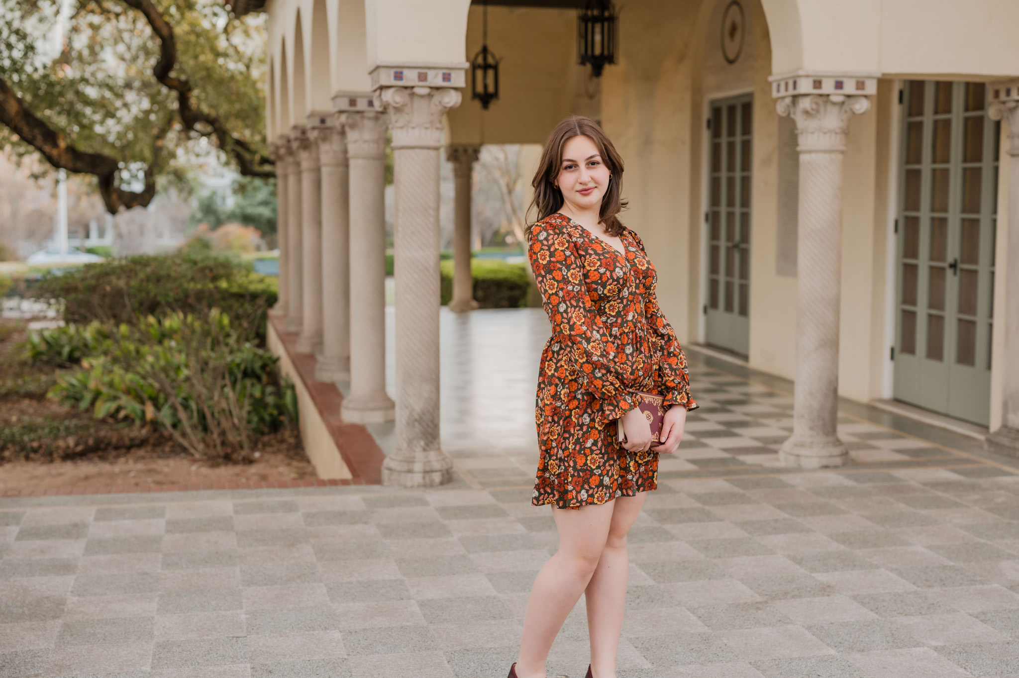 Senior portrait of a girl in a floral dress. She holds a novel and is standing on the back patio of Landa Library in San Antonio.