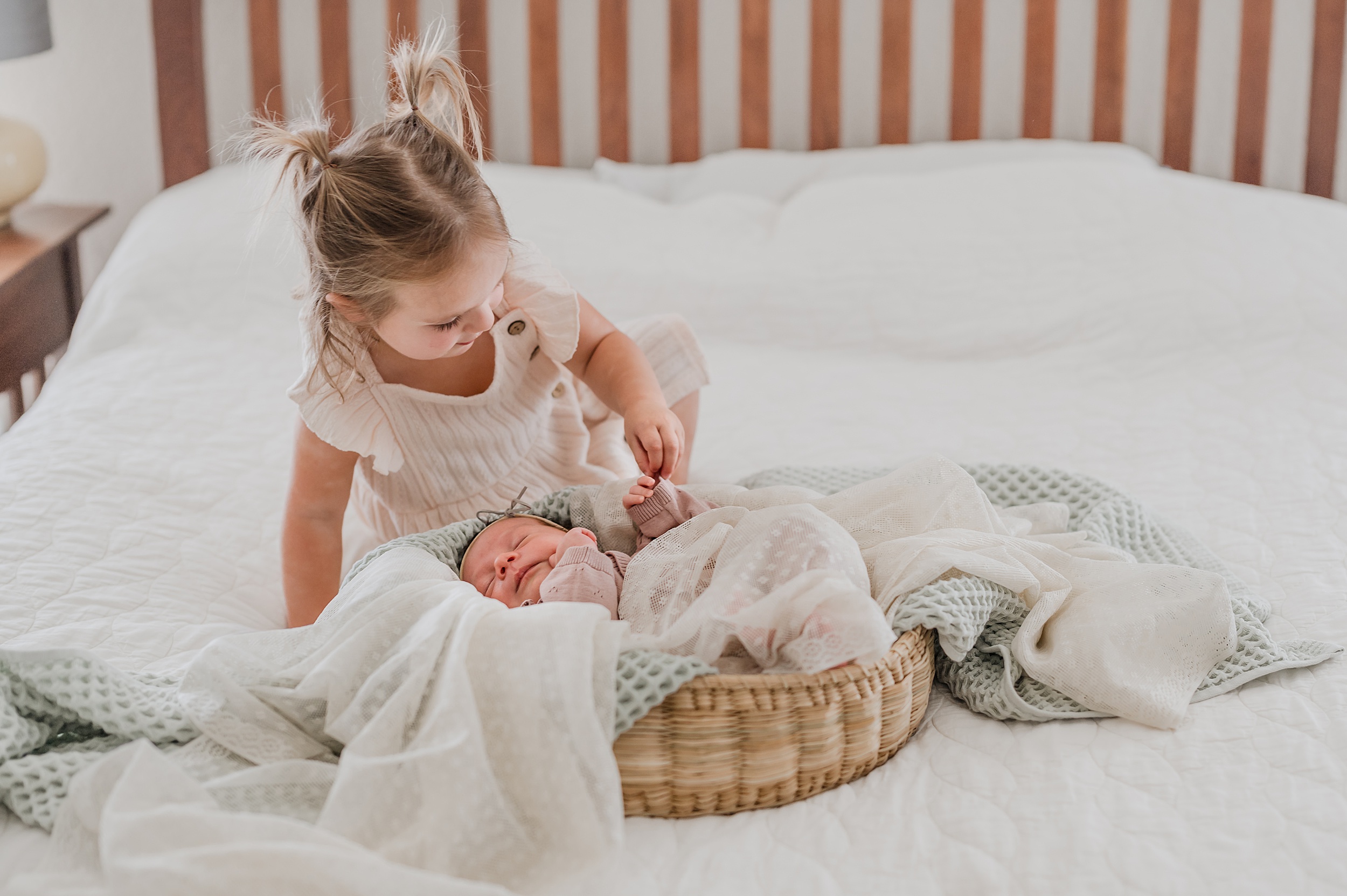 A newborn baby sleeps in a basket full of blankets while her toddler big sister holds her hand on a bed