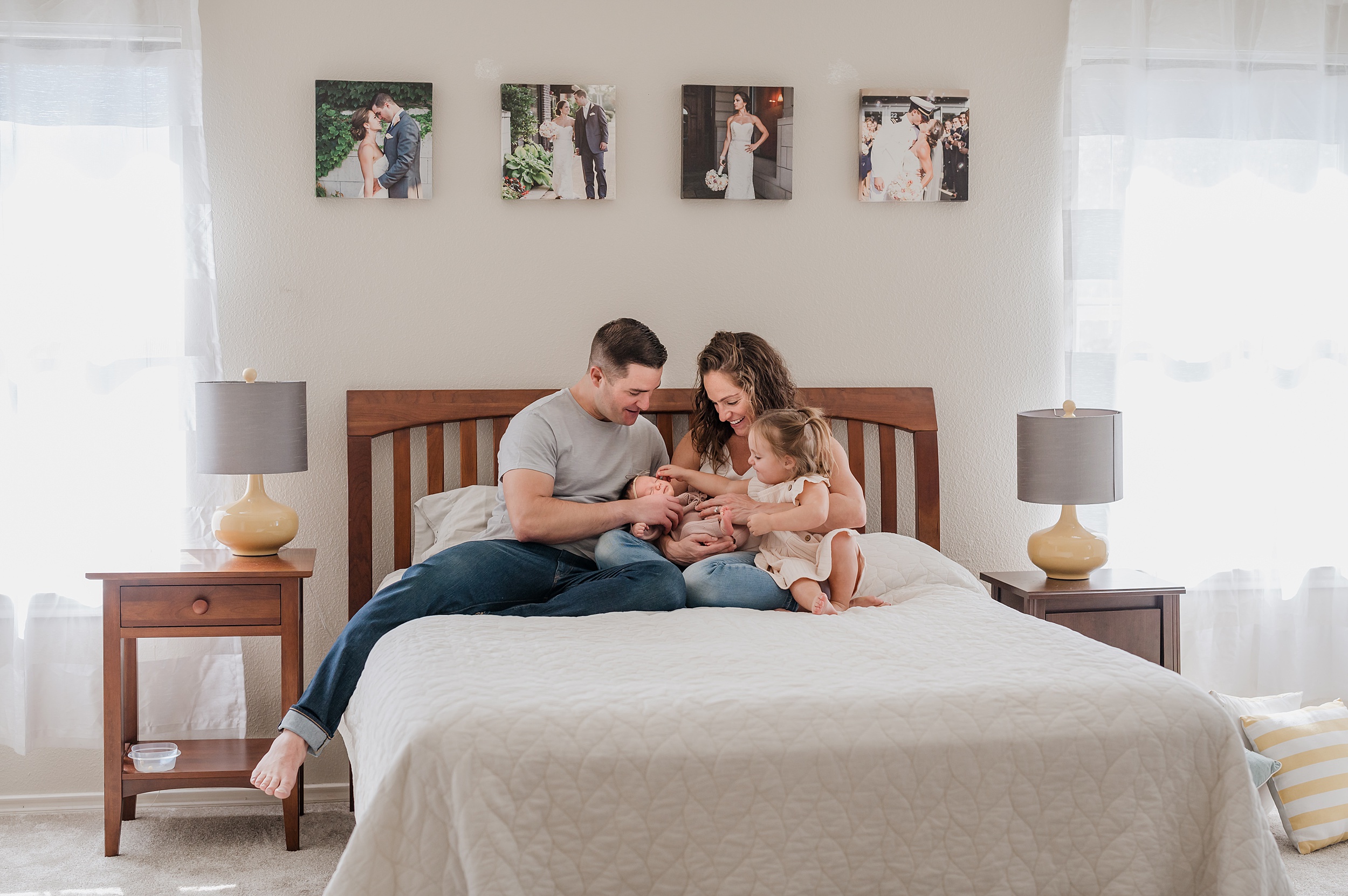 Mom and dad sit on a bed with their newborn baby girl in their laps while their toddler daughter reaches in