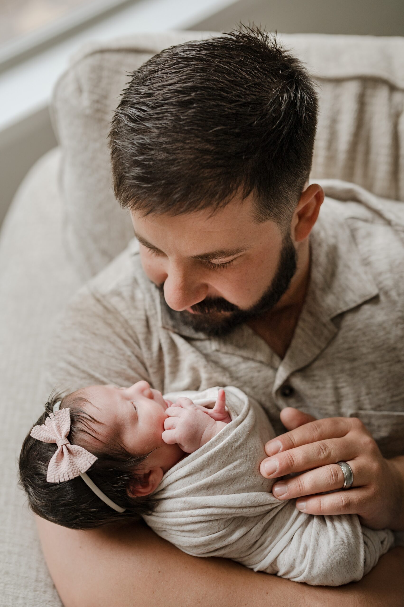 A new dad sits in a large grey chair while holding and looking down at his newborn daughter in his arms