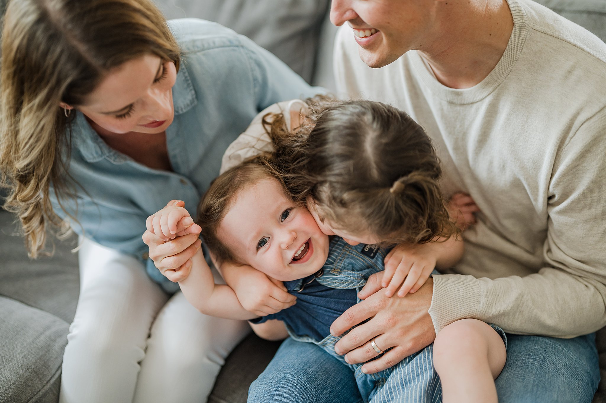 A toddler boy in blue overalls gets tickled by his family while sitting on a couch