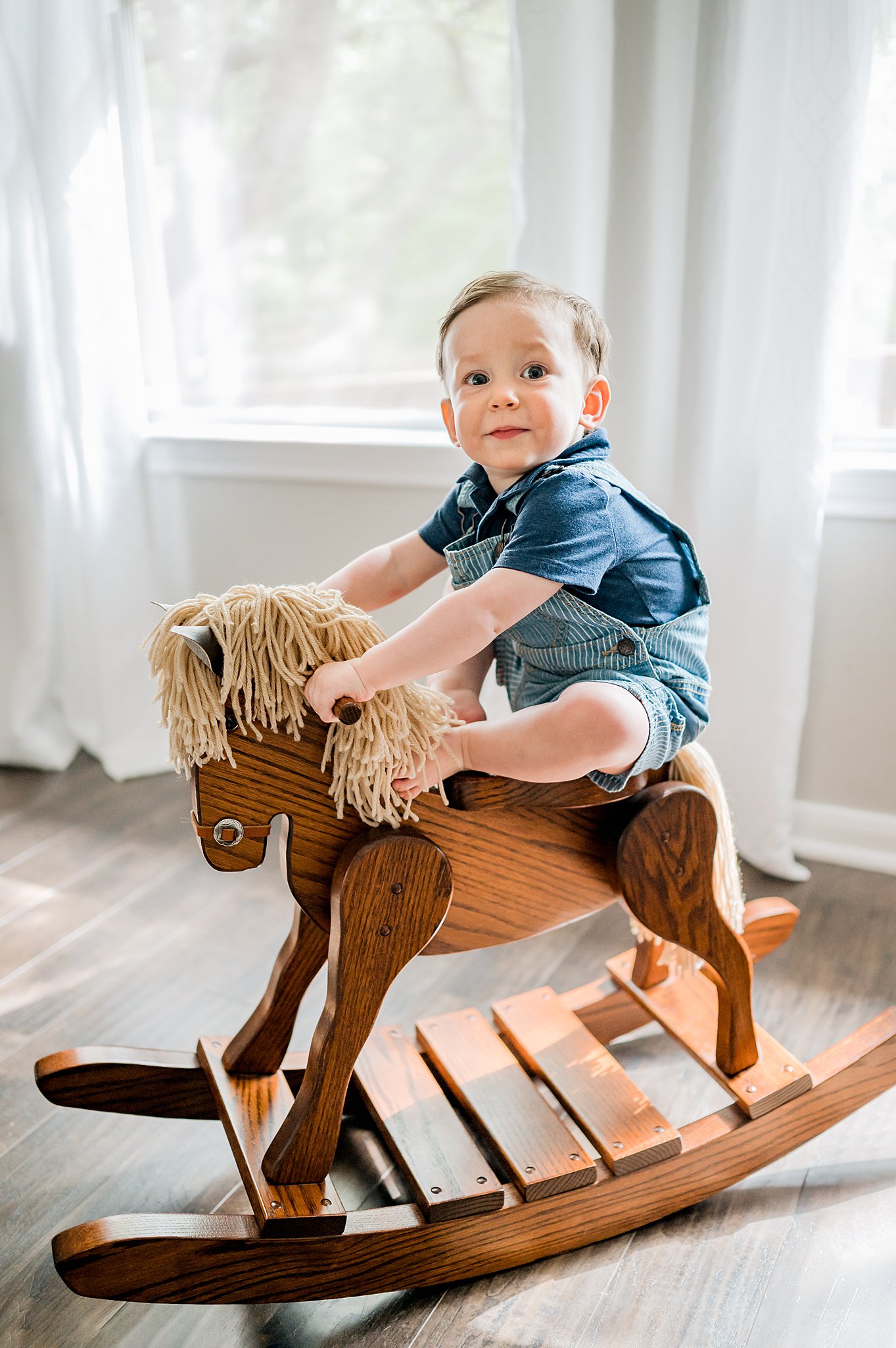 A young boy in blue overalls plays on a wooden rocking horse in front of large windows san antonio toy stores