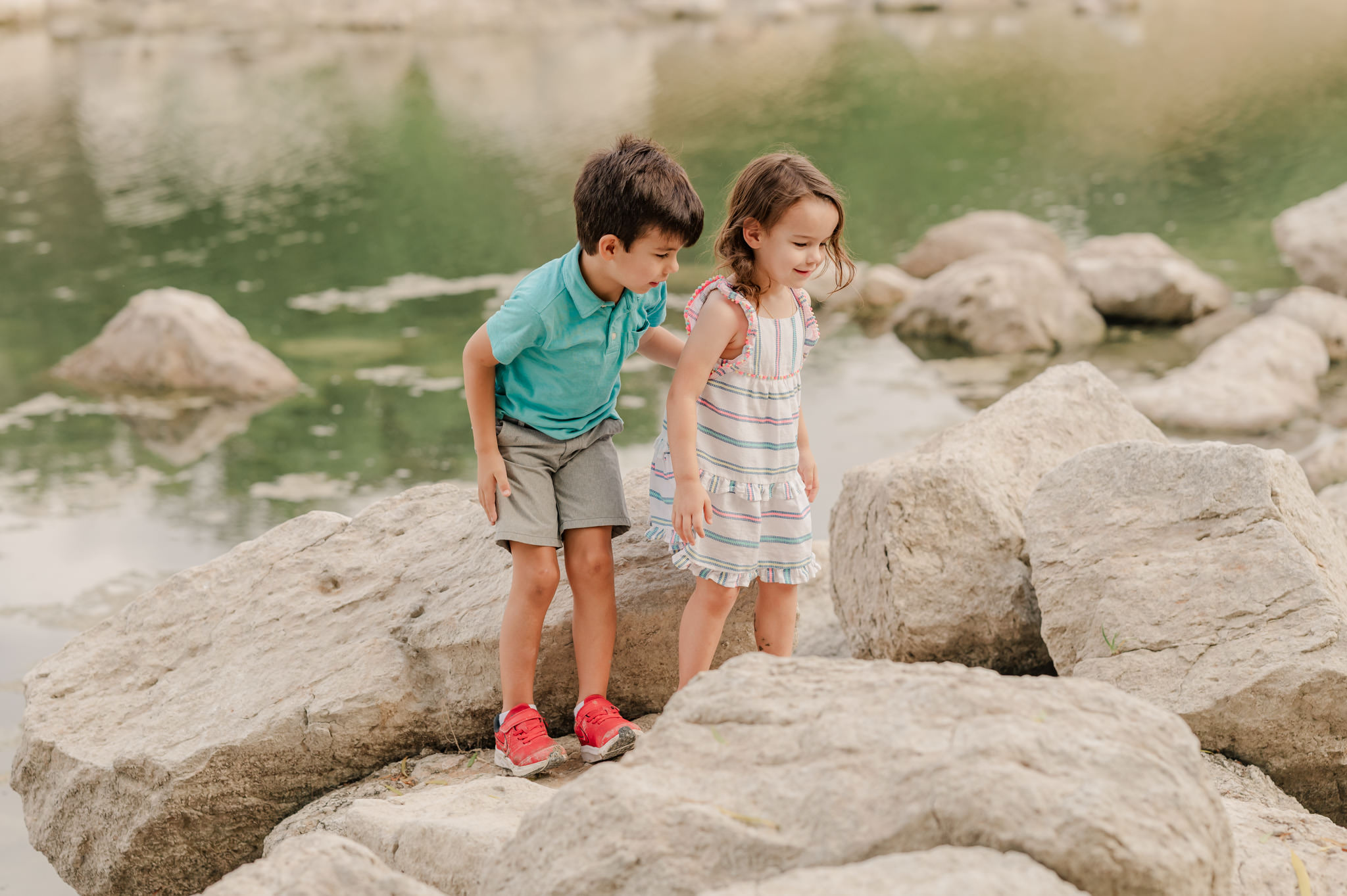 Boy and girl climb on rocks by the water. He's protectively holding her arm. 