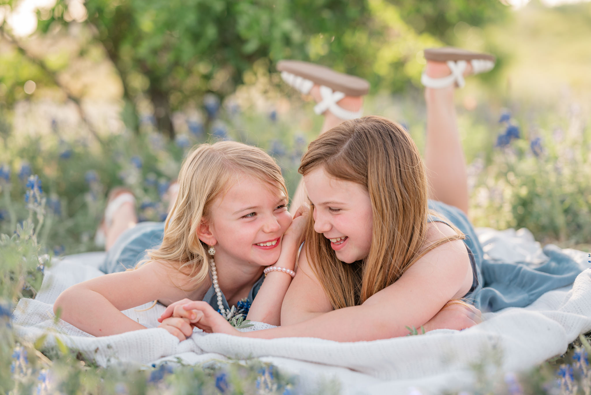 Two sisters whisper and laugh on a blanket among bluebonnets.