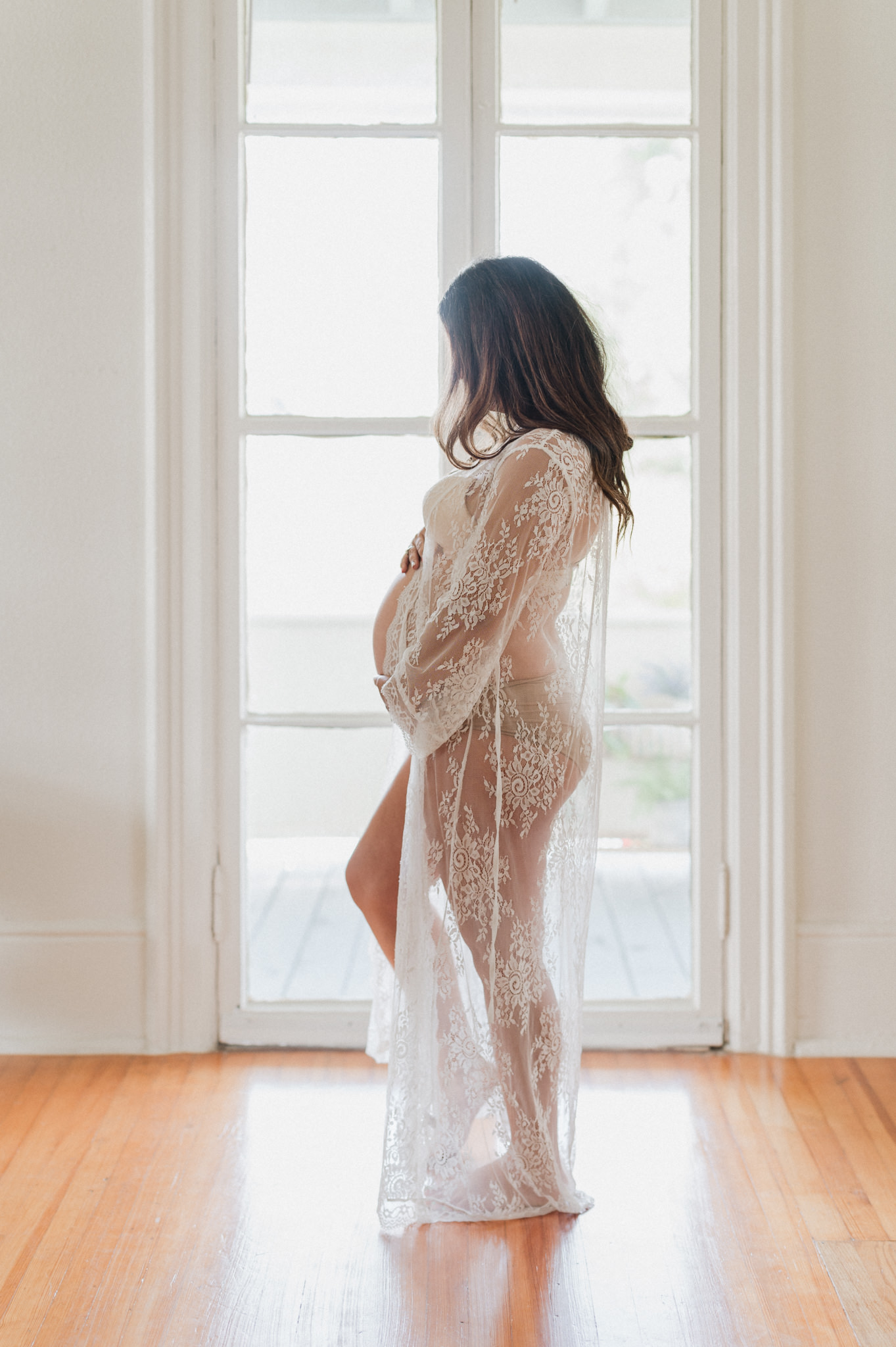 A mother-to-be in a lace maternity gown stands in front of a window looking down at her bump acacia obgyn