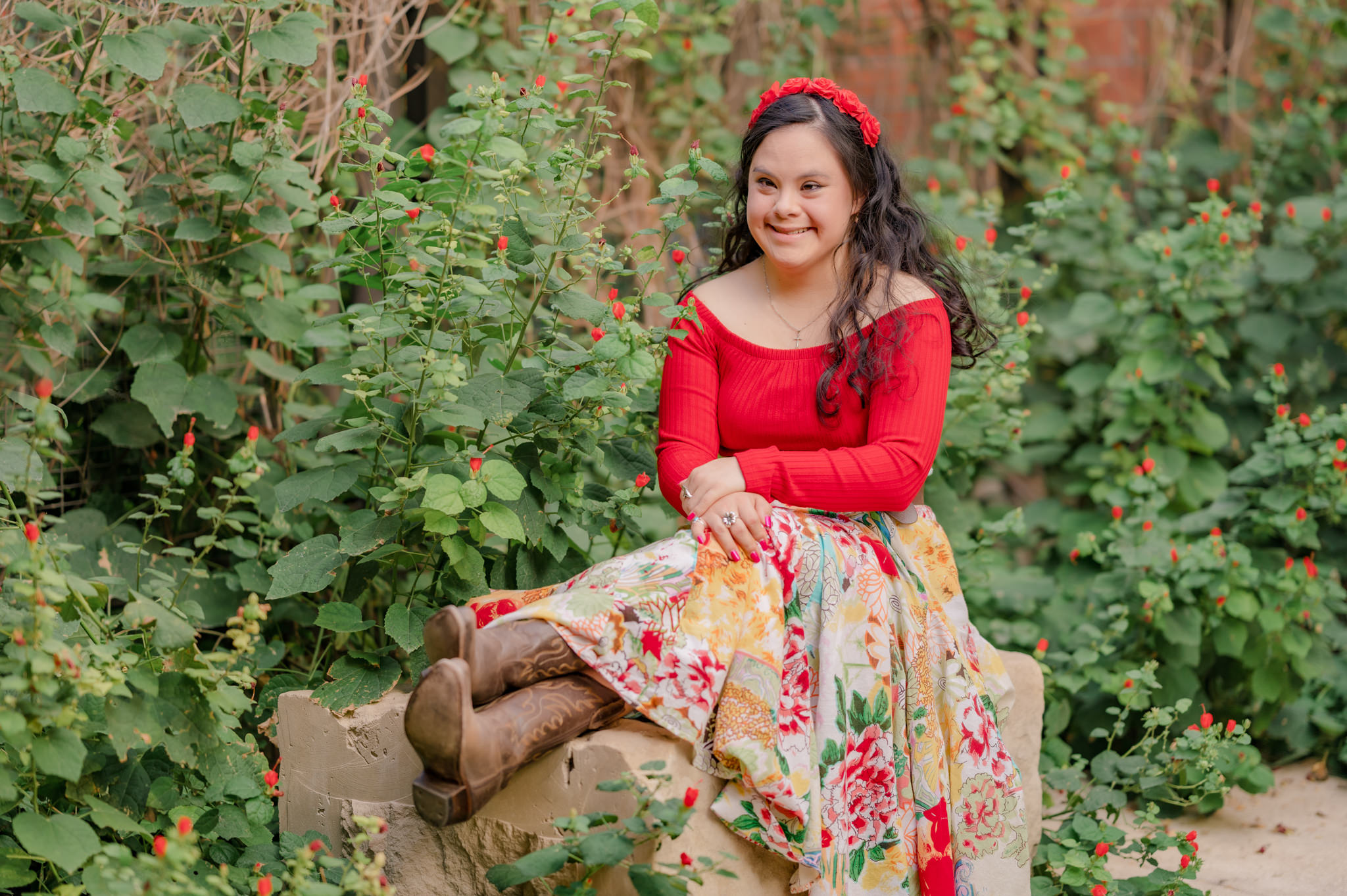 A young woman with Down syndrome looks stunning in a floral skirt and a bright red top amongst a wall of red flowers. Image by San Antonio inclusive photographer Cassey Golden.