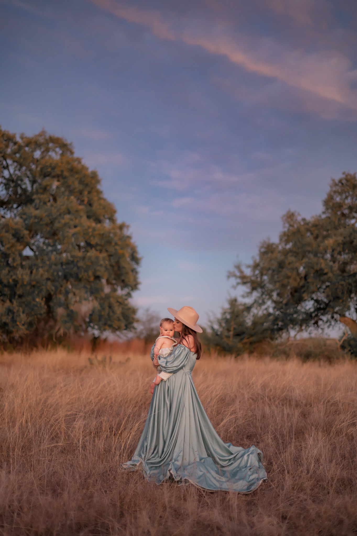 A woman in a flowing blue dress kisses her baby boy on the temple. They stand in a grassy field just after sunset at Joshua Springs Park and Preserve. Image shot by Cassey Golden Photography.