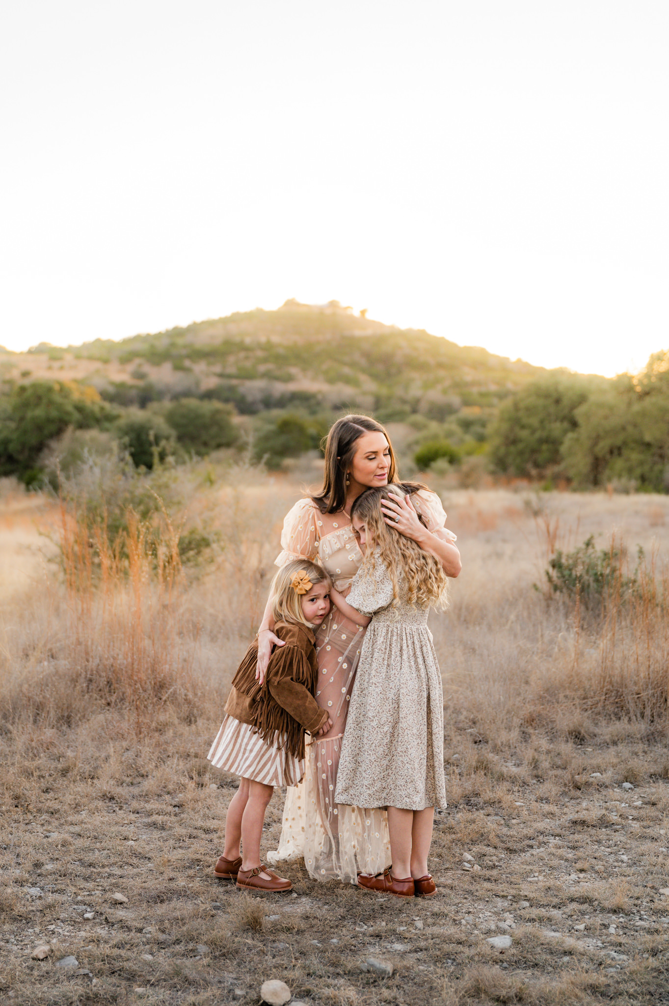 A mom hugs her daughters as the sunset illuminates the hill country view behind them. Picture taken by San Antonio family photographer Cassey Golden at Joshua Springs Park and Preserve. 