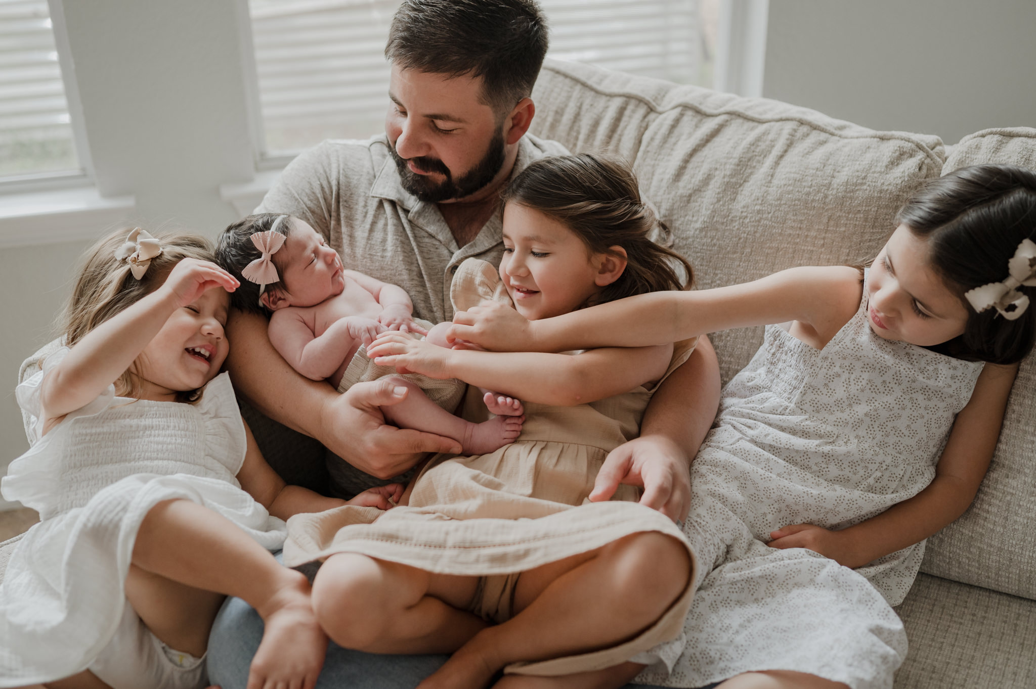 Three sisters sit on a couch playing with their newborn baby sister in dad's arms