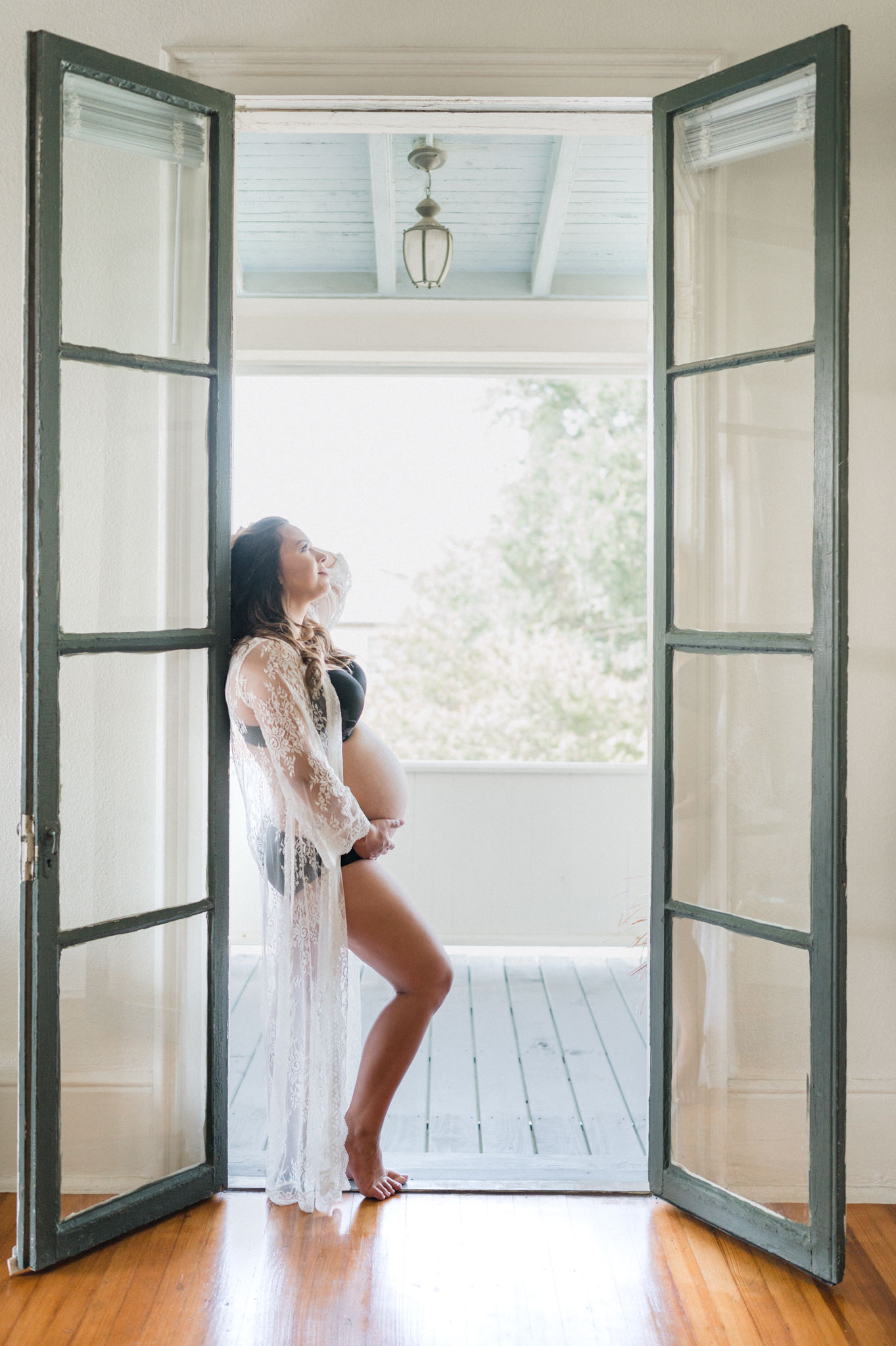 A mom to be in black underwear and a sheer lace coverup stands against a frame of open french doors