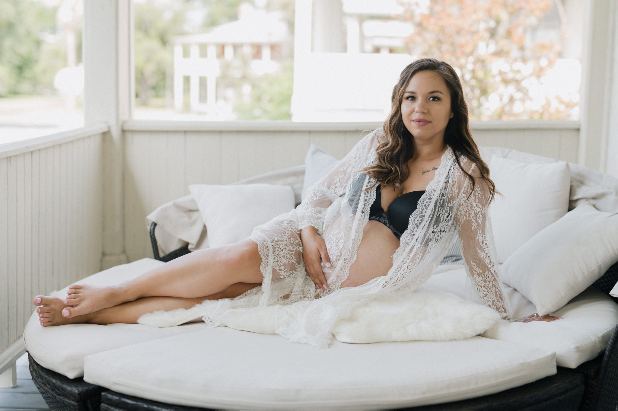 A mother to be in black lingerie and a lace coverup sits across a porch daybed after a san antonio prenatal massage