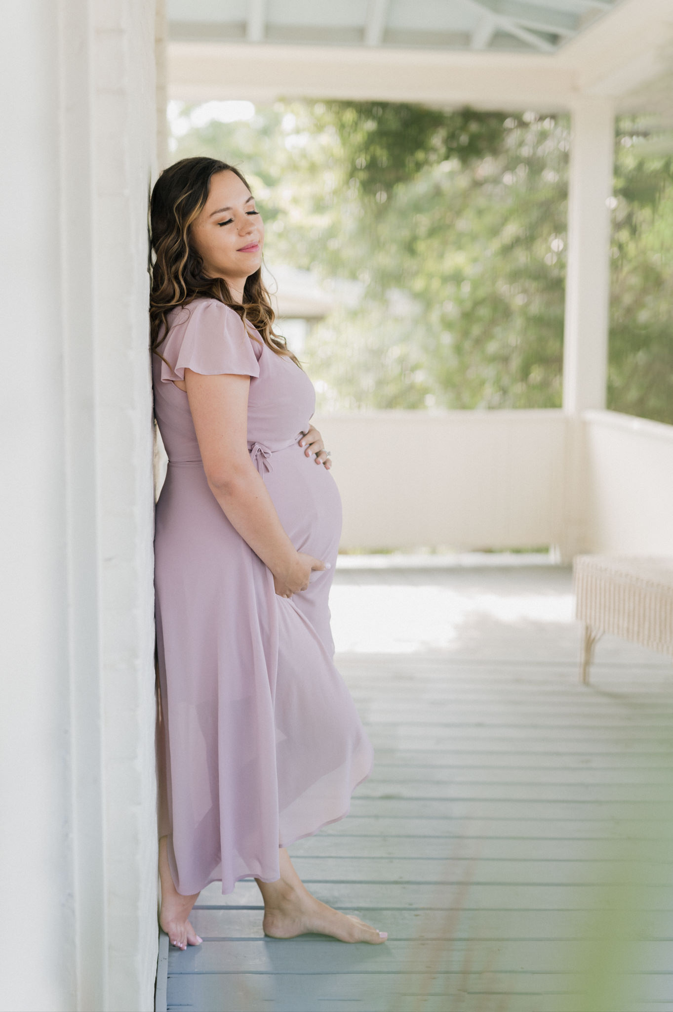 A pregnant mother in a purple maternity gown stands on a porch holding her bump with eyes closed