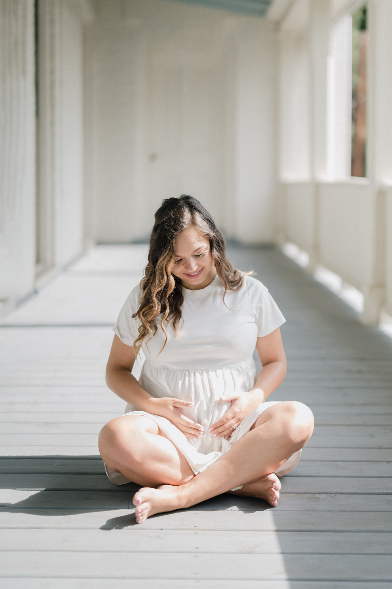 A pregnant mother in a white dress sits crossed legged on a porch in the sun holding her bump